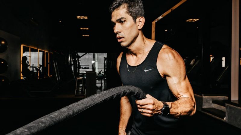 A determined male athlete working out with battle ropes in a dimly lit gym, showcasing strength and fitness.